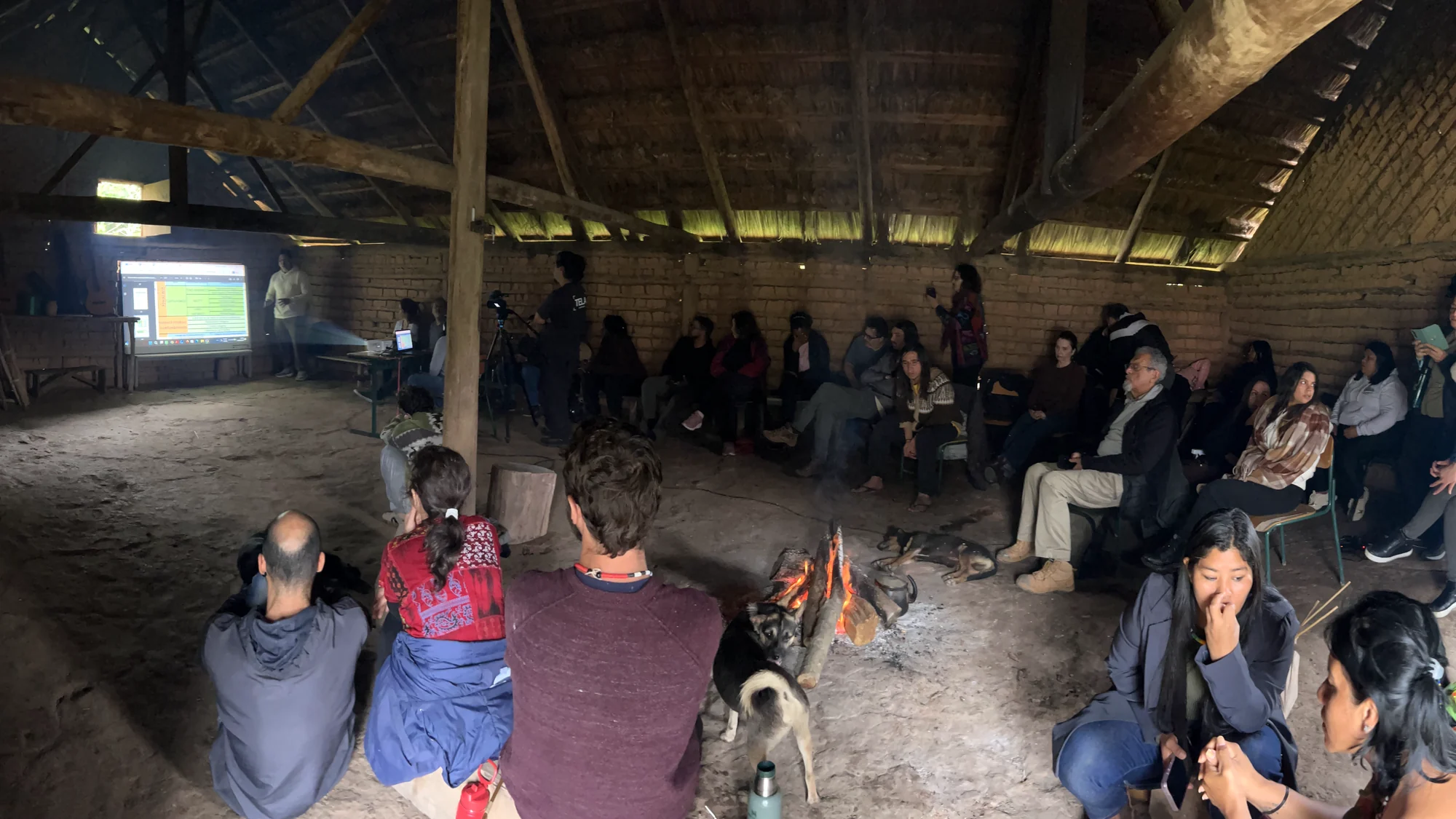 O professor, estudantes e pessoas da aldeia sentados em roda numa casa indígena de barro e madeira, com uma fogueira no chão e uma apresentação projetada na parede