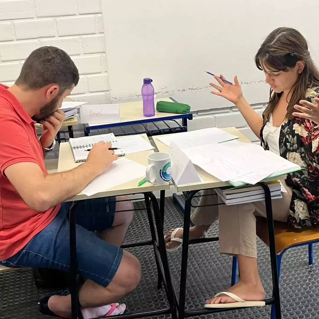 Um estudante escreve num caderno enquanto uma estudante fala, gesticulando, numa mesa cheia de papéis